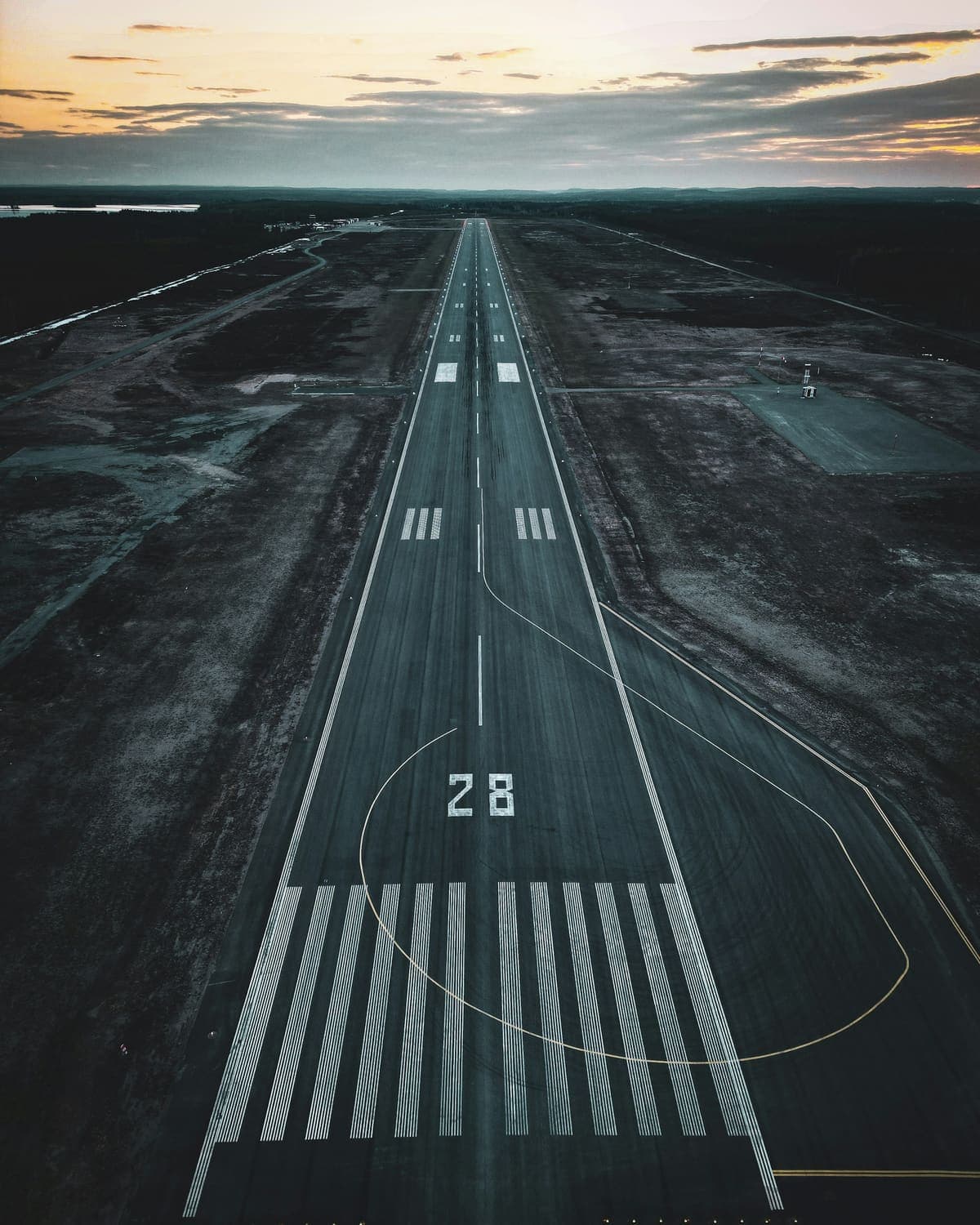 A runway seen from the air along its centreline at dusk, threshold bars and the designator 28 painted across the wet surface