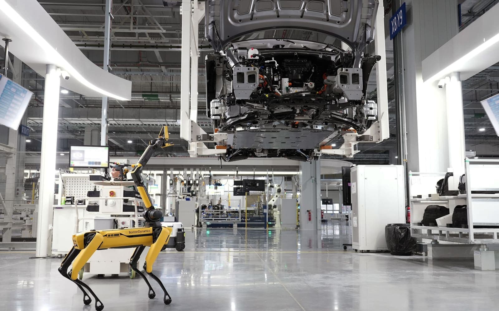 A quadruped inspection robot carrying a manipulator arm standing on the floor of a live vehicle assembly plant, with a body shell on an overhead hoist and an automated guided vehicle behind it