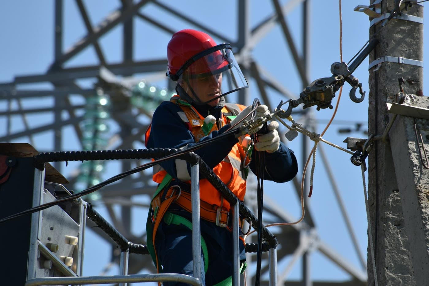 Line worker in a harness and hard hat working on conductor hardware beside a transmission tower