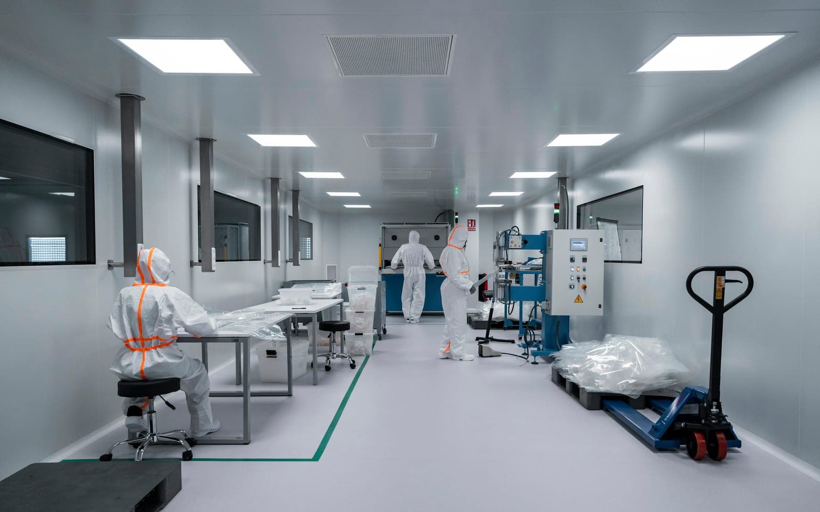 Three gowned operators working in a cleanroom corridor, one seated at a bench, one at the control panel of a filling machine and one at a laminar flow cabinet