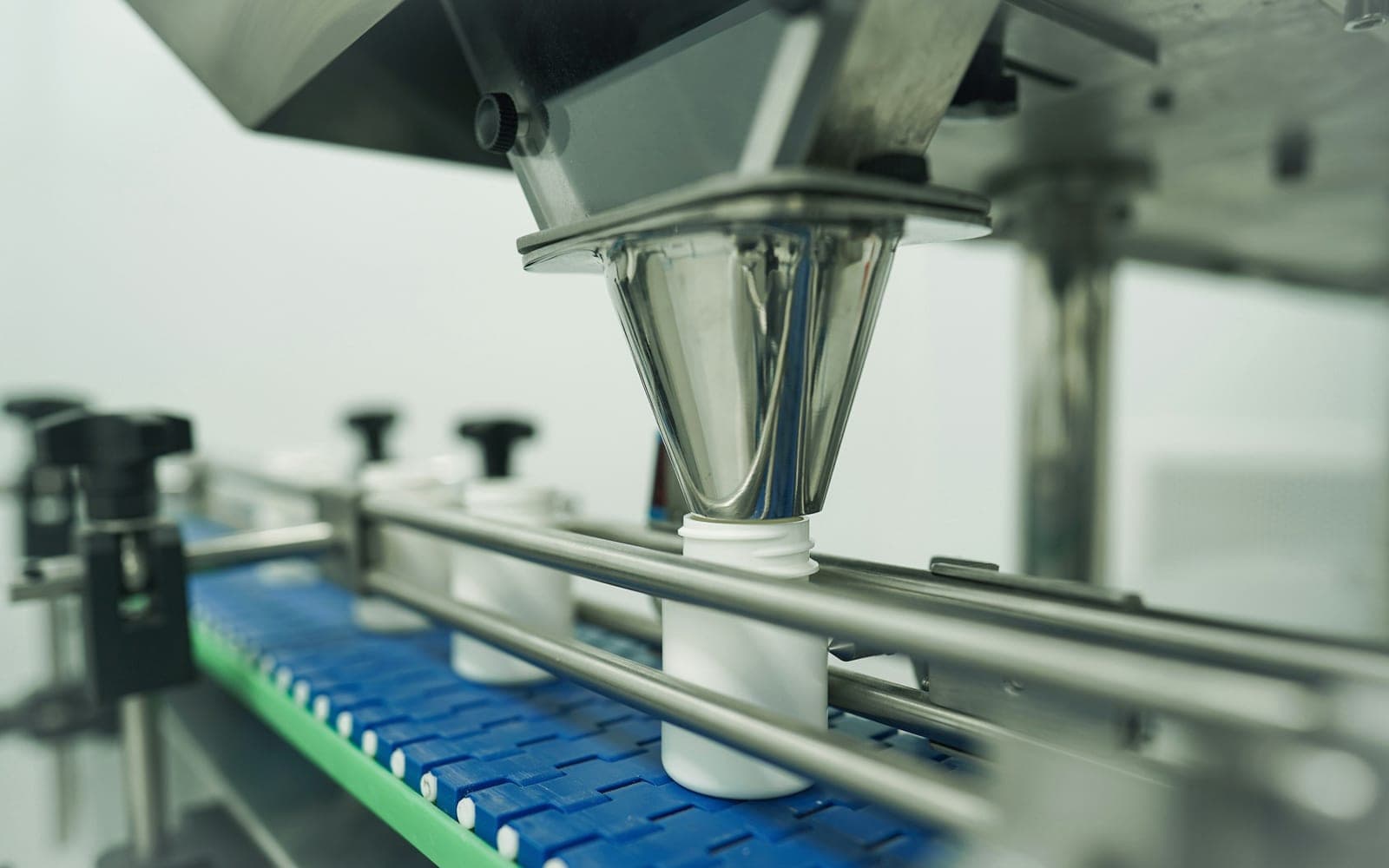 A stainless filling head lowered over a white bottle travelling on the blue chain conveyor of a packaging line