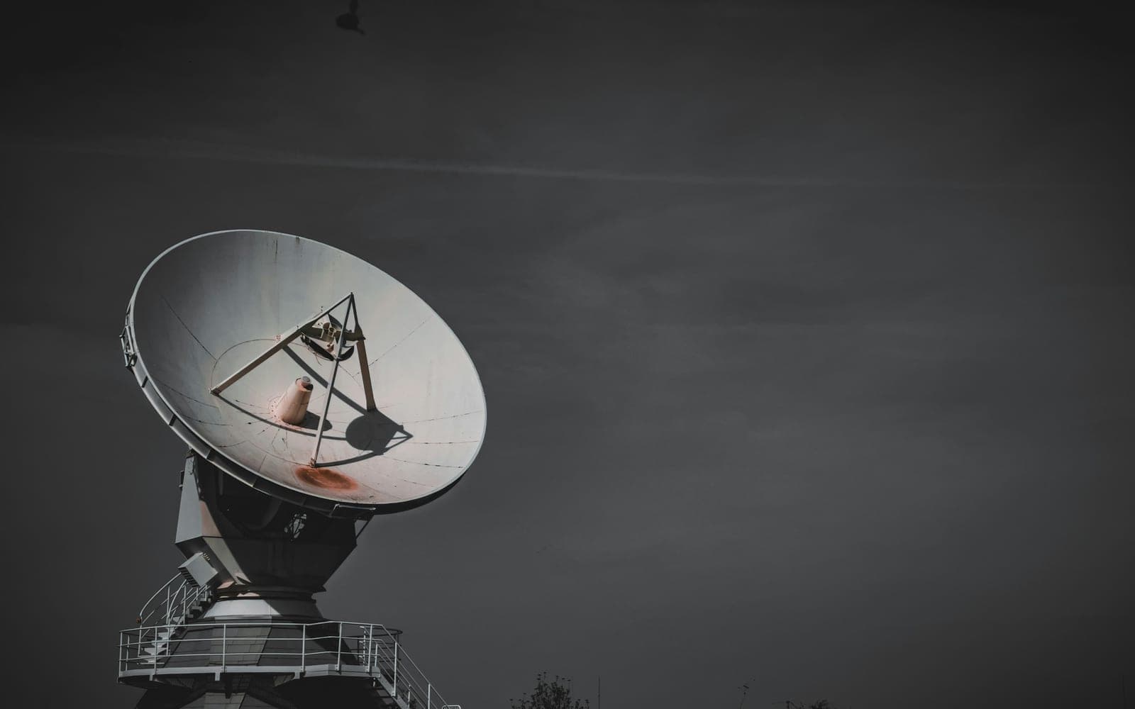 A large steerable communications antenna on a concrete pedestal against a dark overcast sky, its feed assembly and access stair picked out in low light