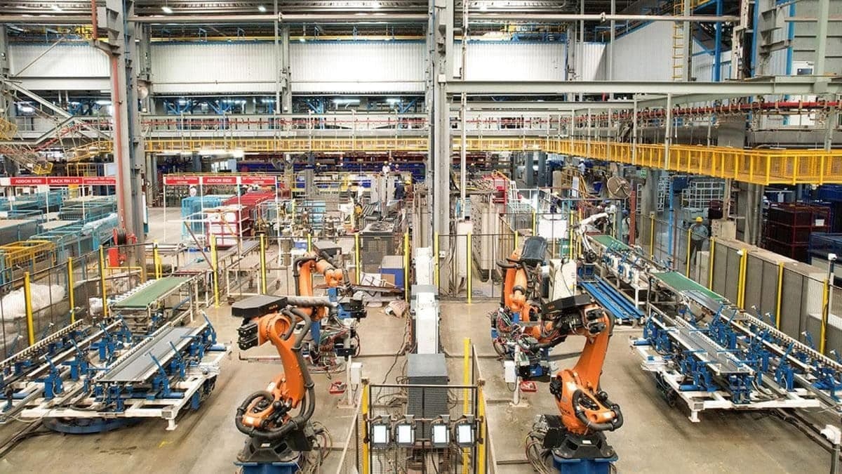 An elevated view of an automated production floor, with two orange articulated robots working between conveyor lines under an overhead crane gantry
