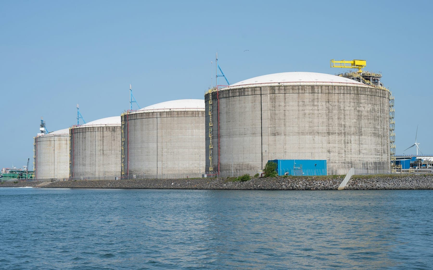 Four full containment liquefied natural gas storage tanks on a waterfront terminal, with jetty gantries and process pipework running behind them